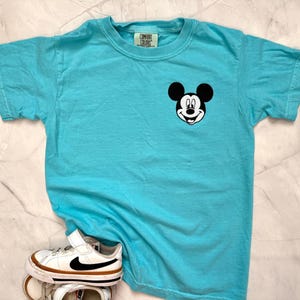 May include: A turquoise Comfort Colors t-shirt featuring a Mickey Mouse graphic on the chest. The shirt is paired with white and brown sneakers with a black swoosh. The t-shirt has a crew neck and short sleeves.