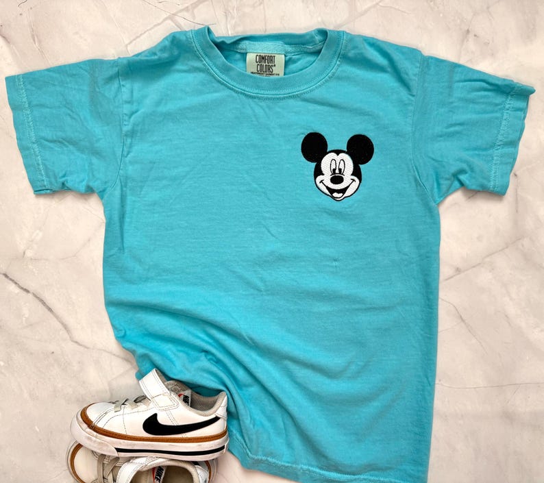 May include: A turquoise Comfort Colors t-shirt featuring a Mickey Mouse graphic on the chest. The shirt is paired with white and brown sneakers with a black swoosh. The t-shirt has a crew neck and short sleeves.