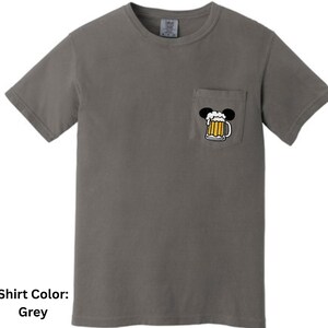 May include: A grey short-sleeved t-shirt with a crew neckline and a single chest pocket. The pocket features a graphic of a beer mug with a foamy head, topped with two black circular ears, creating a silhouette resembling a mouse head shape.