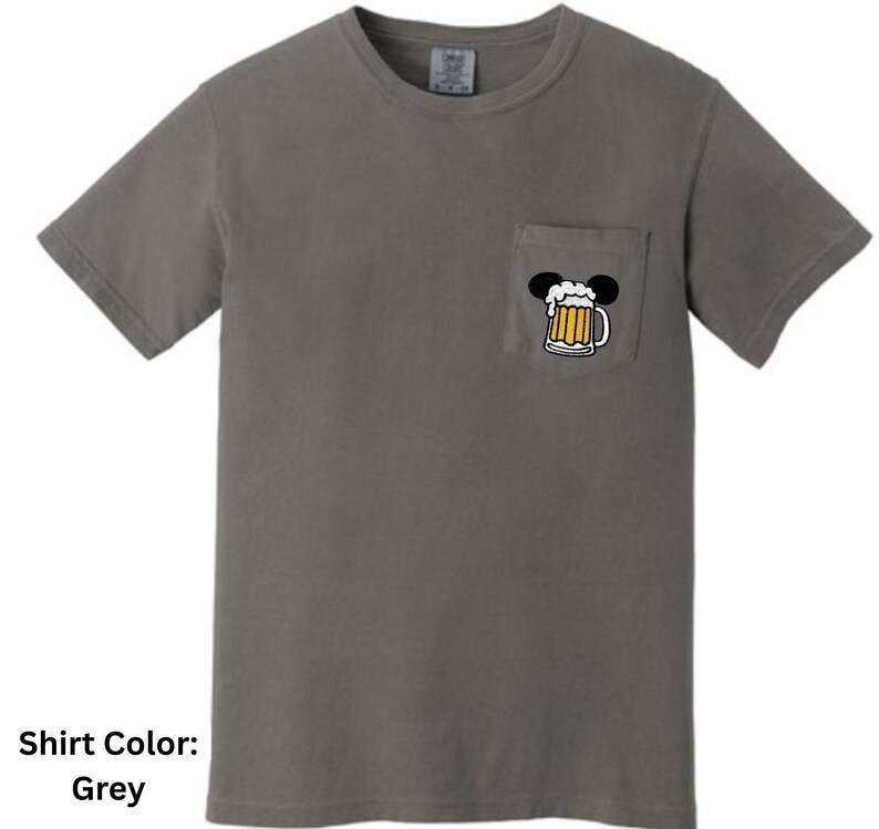 May include: A grey short-sleeved t-shirt with a crew neckline and a single chest pocket. The pocket features a graphic of a beer mug with a foamy head, topped with two black circular ears, creating a silhouette resembling a mouse head shape.