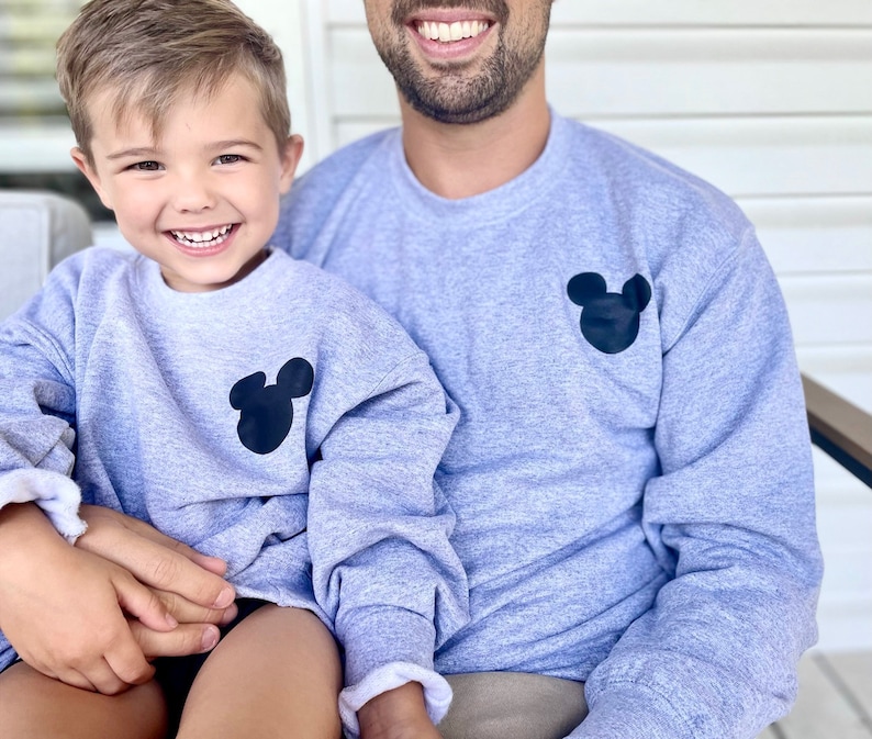 May include: A father and son wearing matching gray sweatshirts with a black Mickey Mouse silhouette on the front.