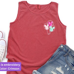 May include: Crimson tank top with embroidered Mickey Mouse balloon design. The sleeveless shirt is paired with denim shorts and white sneakers. The design features three balloons in pink, green, and pink.