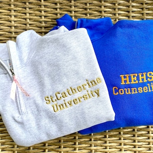 May include: Two gray and blue sweatshirts with embroidered logos. The gray sweatshirt has "St. Catherine University" embroidered in gold. The blue sweatshirt has "HEHS Counseling" embroidered in gold.
