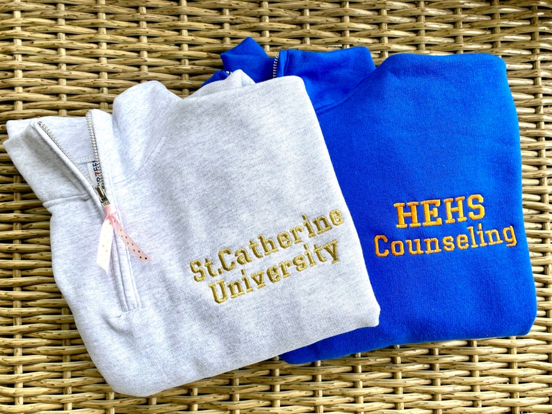 May include: Two gray and blue sweatshirts with embroidered logos. The gray sweatshirt has "St. Catherine University" embroidered in gold. The blue sweatshirt has "HEHS Counseling" embroidered in gold.