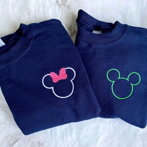 Op de afbeelding: Twee marineblauwe sweatshirts met geborduurde Mickey Mouse en Minnie Mouse ontwerpen. E&eacute;n sweatshirt heeft een groene Mickey Mouse omtrek, de andere een roze strik op het Minnie Mouse ontwerp. Beide sweatshirts hebben een ronde hals.