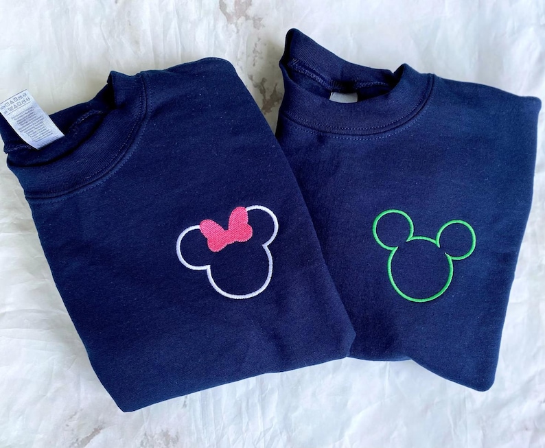 Op de afbeelding: Twee marineblauwe sweatshirts met geborduurde Mickey Mouse en Minnie Mouse ontwerpen. E&eacute;n sweatshirt heeft een groene Mickey Mouse omtrek, de andere een roze strik op het Minnie Mouse ontwerp. Beide sweatshirts hebben een ronde hals.