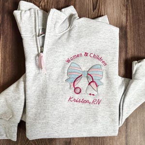 以下が含まれることがあります： 刺繍デザインが施されたライトグレーのクォータージップスウェットシャツ。デザインには、リボン、聴診器、「Women & Children」と「Kristen, RN」の文字がピンクとブルーの糸で刺繍されています。ジッパーにはピンクのプルが付いています。