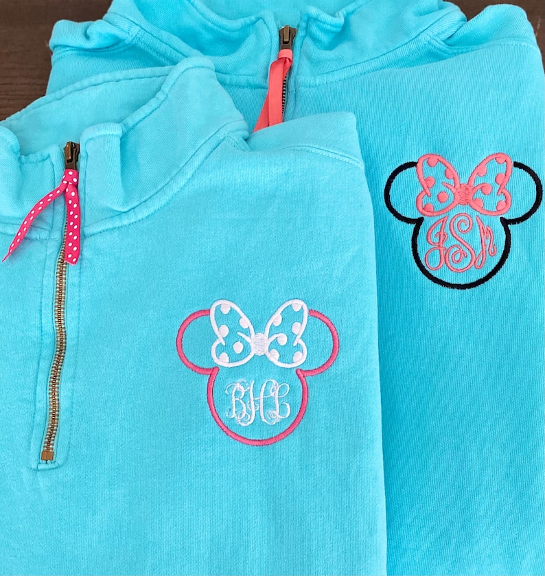 May include: Two turquoise blue quarter-zip sweatshirts with embroidered Minnie Mouse ears and bows. One sweatshirt has the monogram "BHL" and the other has the monogram "JSM".
