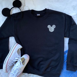 May include: Black sweatshirt with a white Mickey Mouse silhouette made of small white dots. The sweatshirt is laying on a white surface with a pair of white sneakers and a black headband with Mickey Mouse ears.