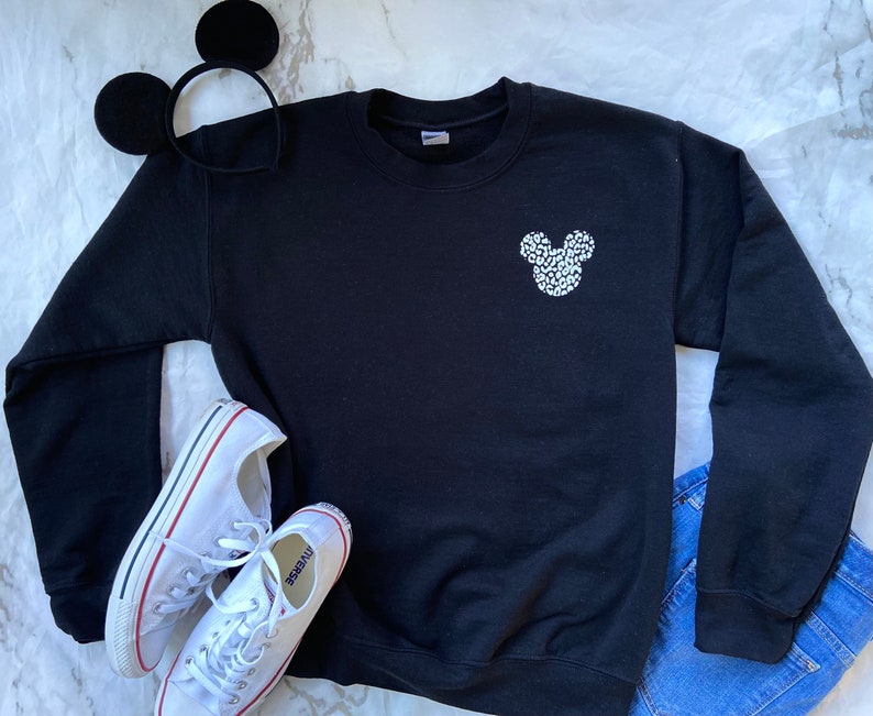 May include: Black sweatshirt with a white Mickey Mouse silhouette made of small white dots. The sweatshirt is laying on a white surface with a pair of white sneakers and a black headband with Mickey Mouse ears.