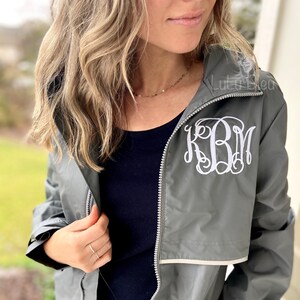 May include: A gray hooded jacket with a white monogram "KBM" embroidered on the front. The jacket has a zipper closure and a white trim around the hood.