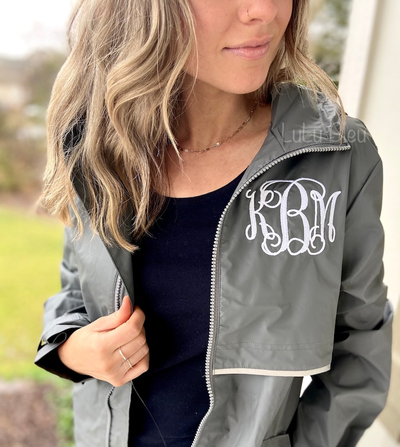 May include: A gray hooded jacket with a white monogram "KBM" embroidered on the front. The jacket has a zipper closure and a white trim around the hood.