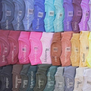 May include: A collection of folded t-shirts arranged in a grid, showcasing a wide variety of colors including true navy, navy, denim, blue jean, washed denim, flo blue, lagoon, chalky mint, violet, berry, neon violet, orchid, red, watermelon, brick, crimson, crunchberry, neon pink, blossom, yam, terracotta, mustard, butter, neon lemon, black, graphite, pepper, espresso, moss, blue spruce, hemp, light green, bay, granite, ivory, and white.