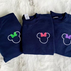Op de afbeelding: Drie marineblauwe sweatshirts met een witte omtrek van een muizenkop en een strik. E&eacute;n strik is groen, &eacute;&eacute;n roze en &eacute;&eacute;n paars. De sweatshirts worden gepresenteerd op een wit oppervlak.