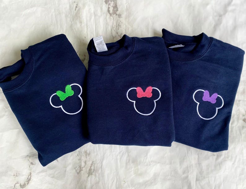 Op de afbeelding: Drie marineblauwe sweatshirts met een witte omtrek van een muizenkop en een strik. E&eacute;n strik is groen, &eacute;&eacute;n roze en &eacute;&eacute;n paars. De sweatshirts worden gepresenteerd op een wit oppervlak.