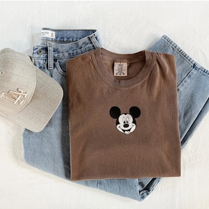May include: A brown crewneck t-shirt featuring a small embroidered Mickey Mouse head graphic on the chest, paired with light blue denim jeans and a beige baseball cap with the letters LA embroidered in white on the front panel.