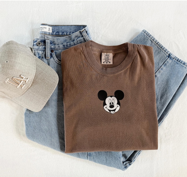 May include: A brown crewneck t-shirt featuring a small embroidered Mickey Mouse head graphic on the chest, paired with light blue denim jeans and a beige baseball cap with the letters LA embroidered in white on the front panel.