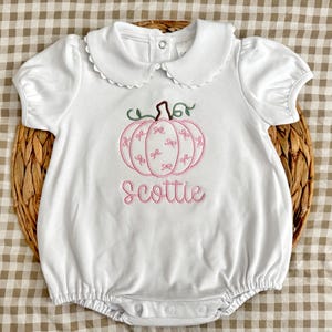 May include: A white short-sleeved bubble romper featuring a scalloped Peter Pan collar and a snap-button closure at the neck. The center displays an embroidered pink pumpkin with small bow patterns and the name Scottie in pink cursive script.