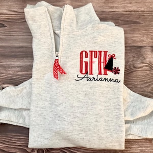 Könnte beinhalten: Ein hellgraues Sweatshirt mit Viertelreißverschluss und einem rot-schwarzen Stickdesign. Das Design umfasst die Initialen "GFH", eine schwarze Glocke und den Namen "Aarianna" in Schreibschrift. Ein rotes Polka-Dot-Band ist am Reißverschluss befestigt.