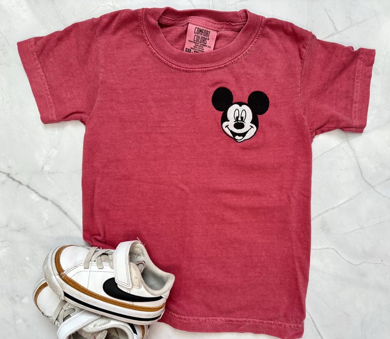 May include: A red t-shirt with a Mickey Mouse patch and a pair of white and gold sneakers. The t-shirt has a crew neck and short sleeves. The sneakers have black accents.