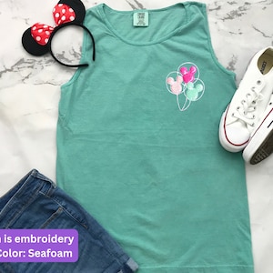 May include: Seafoam green tank top with embroidered Mickey Mouse balloon design. Includes denim shorts, white sneakers, and a Minnie Mouse ear headband with red polka dots. The tank top is a comfortable, casual style.