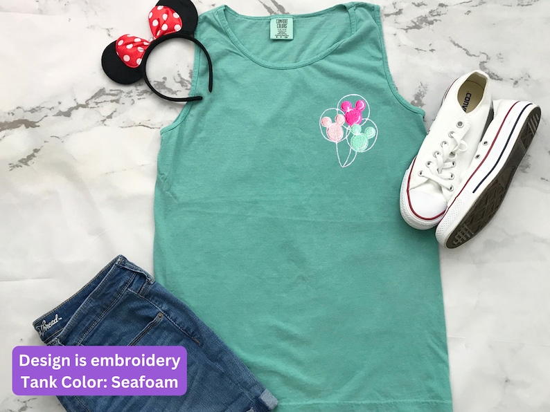 May include: Seafoam green tank top with embroidered Mickey Mouse balloon design. Includes denim shorts, white sneakers, and a Minnie Mouse ear headband with red polka dots. The tank top is a comfortable, casual style.