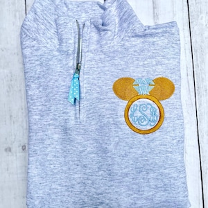 Pu&ograve; includere: Una felpa grigia melange con zip a un quarto, zip di colore blu chiaro e tiretto della zip a pois blu e bianco. La felpa ha un design ricamato di un orecchio di Topolino con un anello di diamanti. L'anello ha il monogramma "LSB" all'interno.