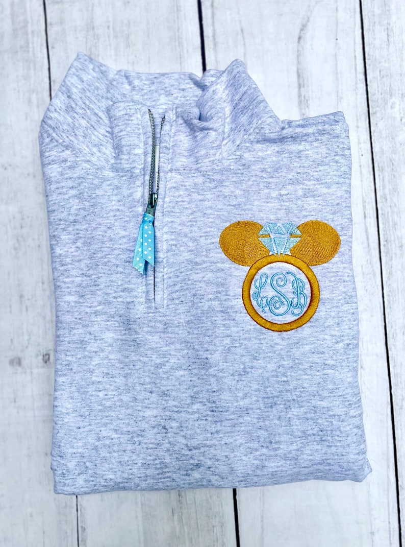 Pu&ograve; includere: Una felpa grigia melange con zip a un quarto, zip di colore blu chiaro e tiretto della zip a pois blu e bianco. La felpa ha un design ricamato di un orecchio di Topolino con un anello di diamanti. L'anello ha il monogramma "LSB" all'interno.