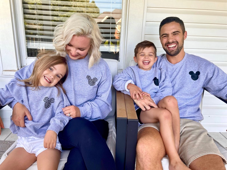 May include: A family of four wearing matching grey sweatshirts with a black Mickey Mouse design. The sweatshirts have a black and white leopard print inside the Mickey Mouse ears. The family are sitting on a porch.