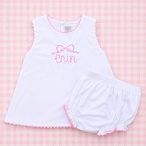 May include: A white sleeveless top and matching bloomer set featuring pink trim along the edges. The top displays an embroidered pink bow above the cursive text enin in pink thread. The set is designed with a simple round neckline and hem.