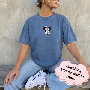 May include: A blue short-sleeved t-shirt featuring a small embroidered graphic of Minnie Mouse on the center chest. The character is depicted with a black and white cow-print bow. The shirt has a crew neckline and a relaxed, casual fit.