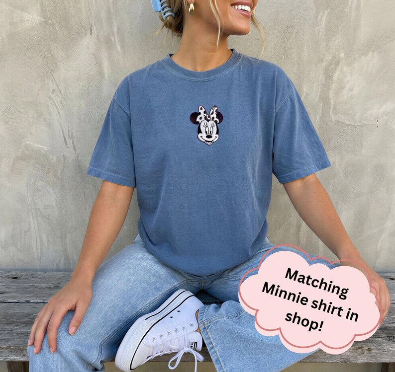 May include: A blue short-sleeved t-shirt featuring a small embroidered graphic of Minnie Mouse on the center chest. The character is depicted with a black and white cow-print bow. The shirt has a crew neckline and a relaxed, casual fit.