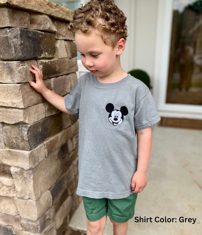 May include: A grey t-shirt featuring a black and white Mickey Mouse graphic on the chest. The shirt is paired with green shorts. The text "Shirt Color: Grey" is visible at the bottom of the image.