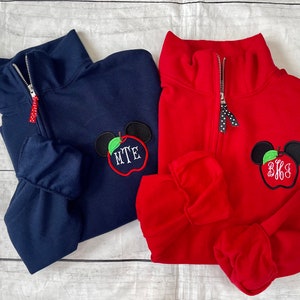 Op de afbeelding: Twee kwart-rits truien met een rood appel en Mickey Mouse oren ontwerp. Een trui is marineblauw met het monogram "MTE" en de andere is rood met het monogram "BKS".