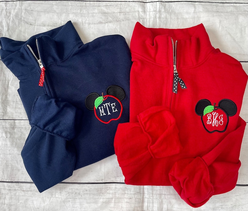 Op de afbeelding: Twee kwart-rits truien met een rood appel en Mickey Mouse oren ontwerp. Een trui is marineblauw met het monogram "MTE" en de andere is rood met het monogram "BKS".