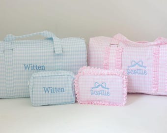 May include: A light blue gingham duffel bag and matching rectangular pouch, both embroidered with the name Witten. Beside them, a pink gingham duffel bag and matching ruffled pouch, both embroidered with the name Scottie and a blue bow icon.