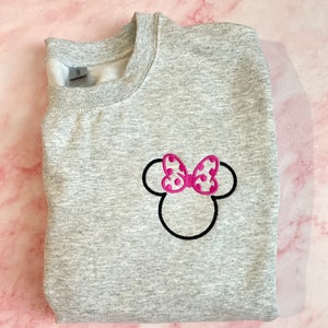 May include: Gray sweatshirt with an embroidered Minnie Mouse head design. The bow is pink with white polka dots.