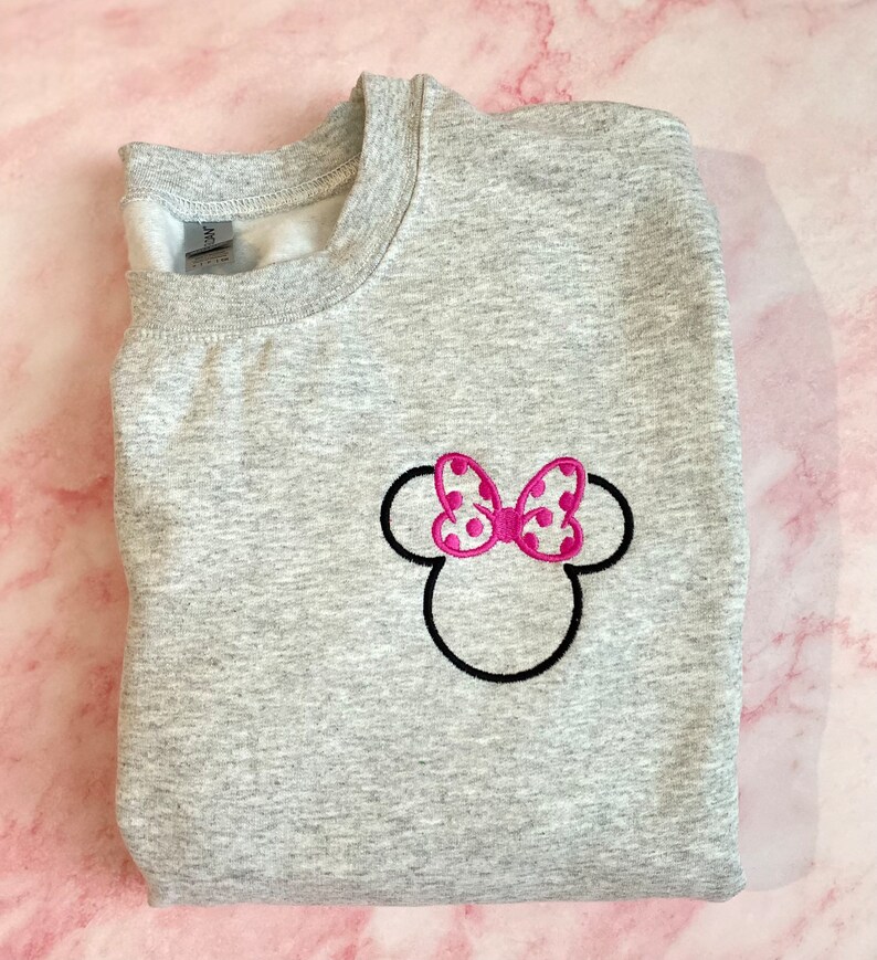 May include: Gray sweatshirt with an embroidered Minnie Mouse head design. The bow is pink with white polka dots.