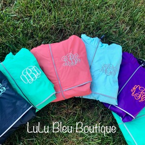 May include: Six different color lightweight jackets with reflective trim and personalized monograms. The jackets are laid out on a bed of green grass. The text "Lulu Bleu Boutique" is visible in the center of the image.