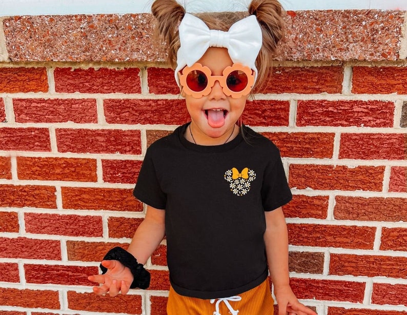 May include: A young girl wearing a black t-shirt with a yellow and white floral Minnie Mouse design, a white bow in her hair, and orange flower-shaped sunglasses. She is standing in front of a red brick wall.