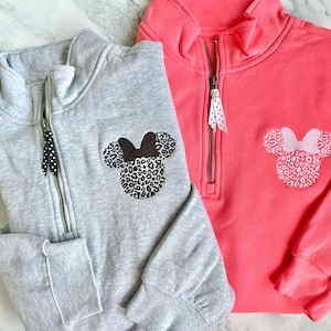 May include: Two sweatshirts with a zipper and a Mickey Mouse head design. One sweatshirt is gray with a black and white Mickey Mouse head and the other is pink with a white Mickey Mouse head.
