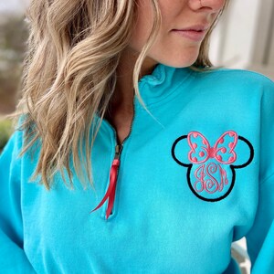 May include: A turquoise blue quarter-zip sweatshirt with a red zipper pull. The sweatshirt features a black and white embroidered design of a mouse head with a pink polka dot bow. The monogram "J.S." is embroidered in black inside the bow.