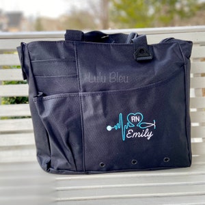 Puede incluir: Bolso tote negro con m&uacute;ltiples bolsillos y una parte superior con cremallera. El bolso tiene un dise&ntilde;o bordado en azul de un coraz&oacute;n con las letras "RN" en su interior, un estetoscopio y una l&iacute;nea de latido del coraz&oacute;n con el nombre "Emily" debajo.