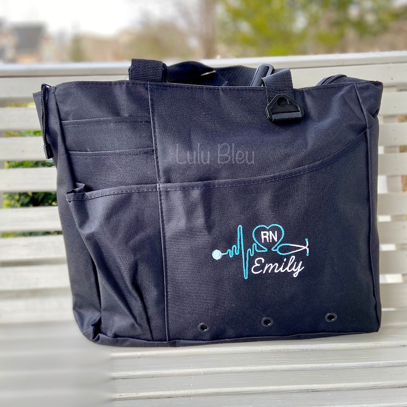 Puede incluir: Bolso tote negro con m&uacute;ltiples bolsillos y una parte superior con cremallera. El bolso tiene un dise&ntilde;o bordado en azul de un coraz&oacute;n con las letras "RN" en su interior, un estetoscopio y una l&iacute;nea de latido del coraz&oacute;n con el nombre "Emily" debajo.