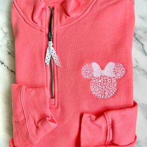 May include: A coral pink quarter-zip sweatshirt with a white embroidered Minnie Mouse head with a leopard print design. The zipper has a white polka dot ribbon pull.