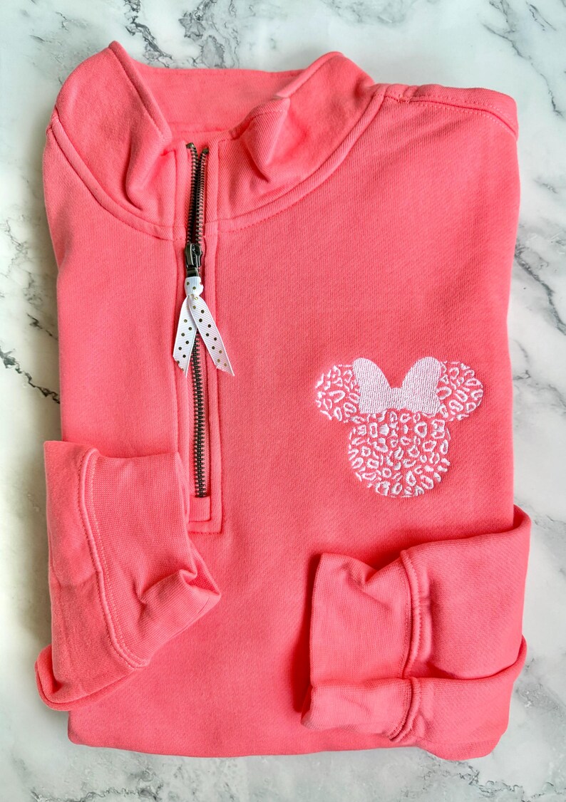 May include: A coral pink quarter-zip sweatshirt with a white embroidered Minnie Mouse head with a leopard print design. The zipper has a white polka dot ribbon pull.