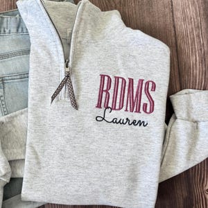 Peut inclure: Un sweat-shirt gris clair à fermeture éclair quart de cercle avec les initiales brodées "RDMS" en bordeaux et le nom "Lauren" en écriture noire. La fermeture éclair a un ruban décoratif. Le sweat-shirt est associé à un jean bleu.