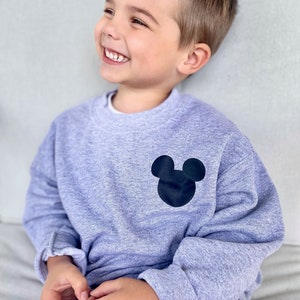May include: A young child wearing a grey sweatshirt with a black Mickey Mouse silhouette on the front. The child is smiling and looking to the right.