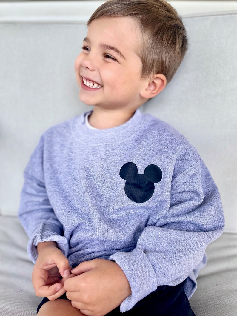 May include: A young child wearing a grey sweatshirt with a black Mickey Mouse silhouette on the front. The child is smiling and looking to the right.