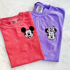 May include: Two Comfort Colors t-shirts, one coral and one purple, each with a cartoon character face embroidered on the chest. The coral shirt features Mickey Mouse, and the purple shirt features Minnie Mouse.